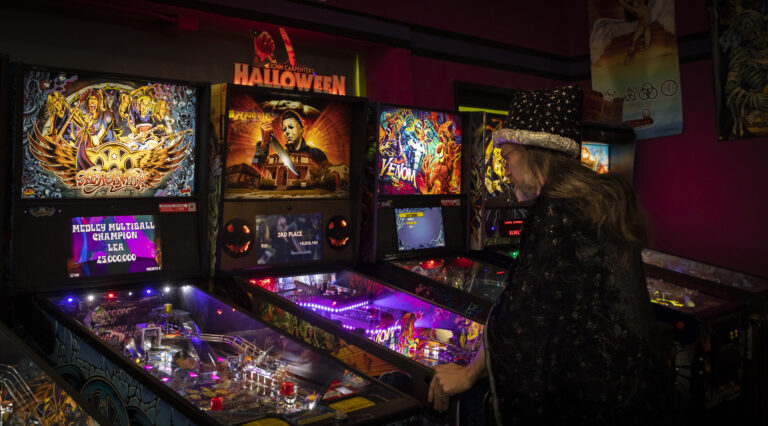A person in a sparkly hat plays pinball in an arcade filled with colorful and illuminated machines featuring horror and superhero themes.