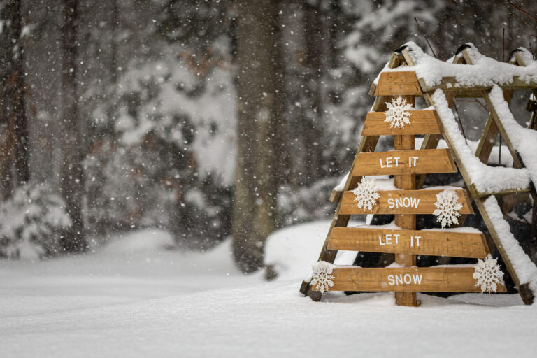 A wooden triangular Christmas tree with the phrase "Let It Snow" spelled out on its planks stands amidst a gentle snowfall.