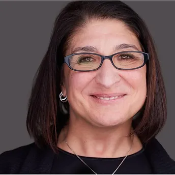A portrait of a woman with medium dark hair and glasses, wearing a black outfit and a subtle smile, set against a neutral gray background.