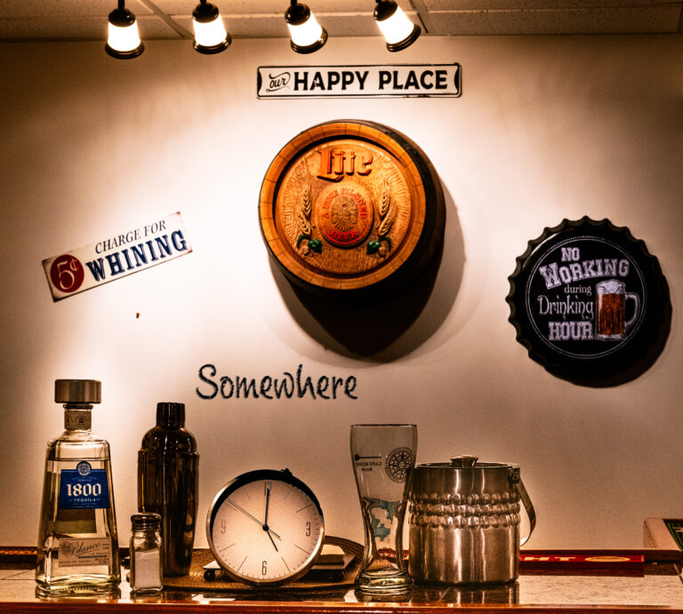 A themed bar wall with various signs, a barrel, beer cap designs, and assorted barware under lights