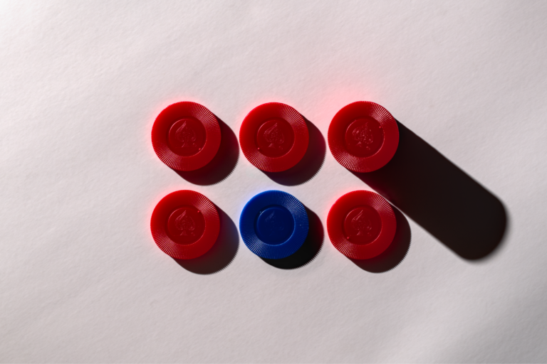 Six red and one blue plastic game tokens arranged symmetrically on a white surface with shadow play